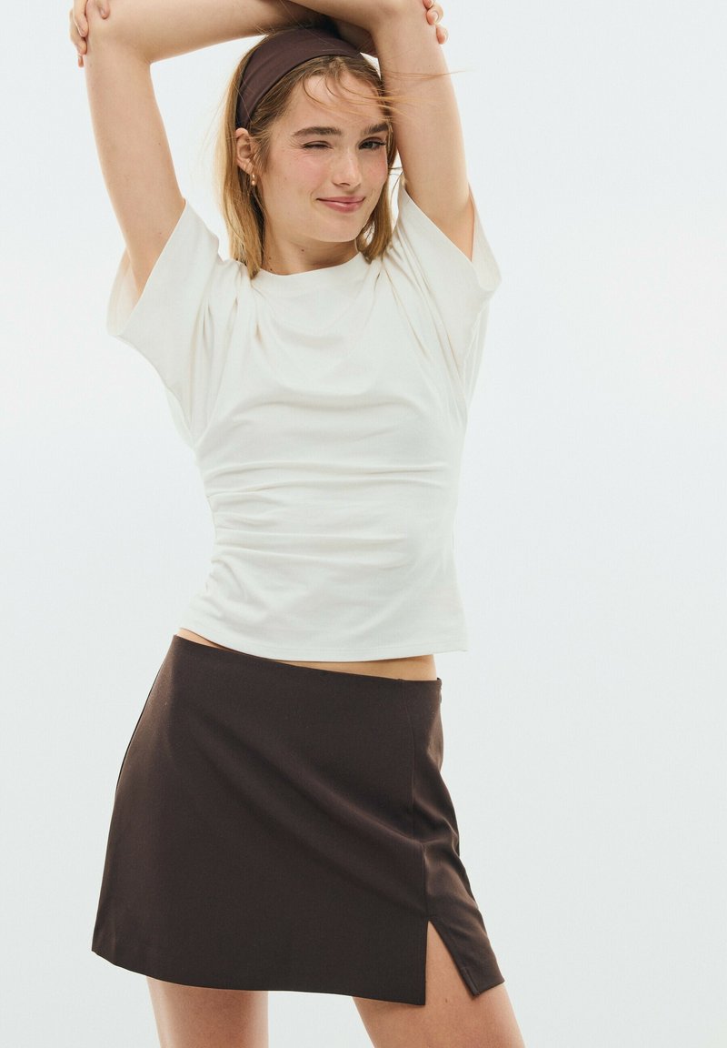 Young woman in white short-sleeve top and dark skirt with side slit stretches arms above head, winking with slight smile.