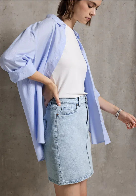 Frau mit hellblauem Jeansrock, weißem Shirt und offenem Hemd vor grauer Wand.