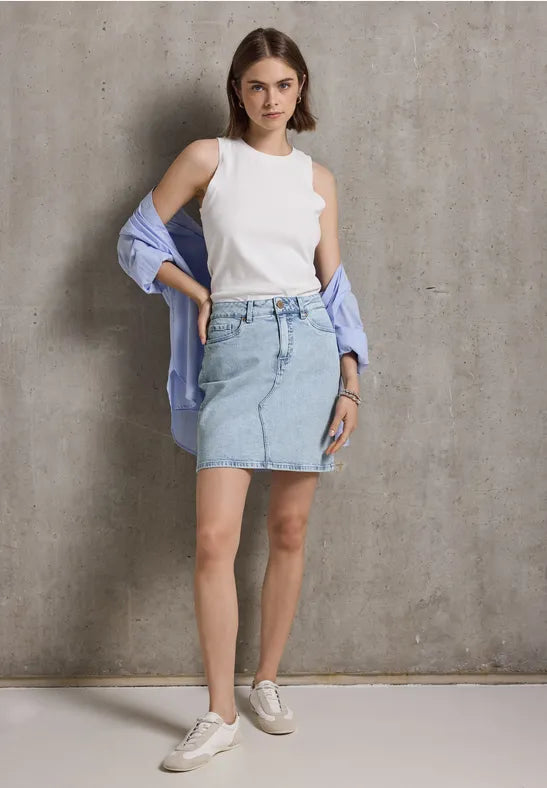 Frau in weißem Top, Jeansrock und Sneakers vor grauer Wand.