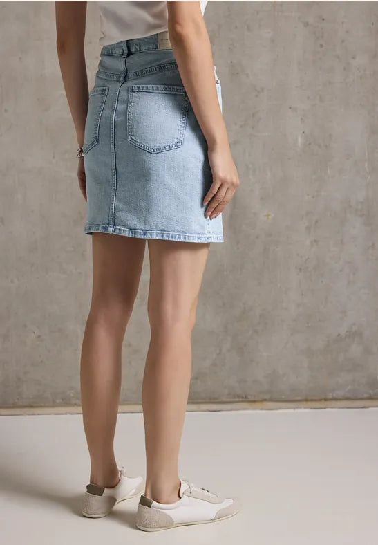 Frau mit Jeansrock und Sneakers steht vor grauer Wand.