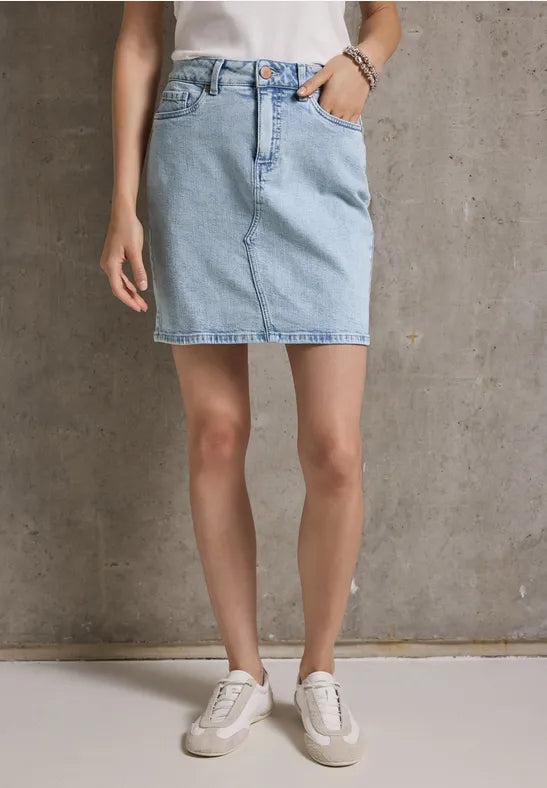 Frau mit Jeansrock, weißem Top und Sneakers vor Betonwand.